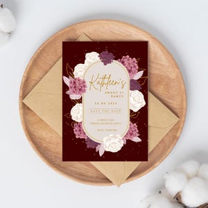 May include: A maroon invitation card with gold text and floral accents, featuring the text "Kathleen's SWEET 17 PARTY" and "SAVE THE DATE". The card is surrounded by white and purple flowers and rests on a wooden plate with a brown envelope.