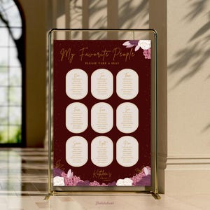 May include: A dark burgundy wedding seating chart sign displayed on a gold metal stand. The sign features the text My Favorite People, Please Take A Seat, and lists nine tables with floral accents in white and purple at the top and bottom.