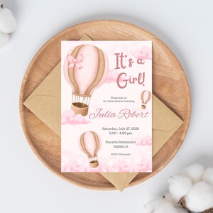 May include: A baby shower invitation with a pink and brown hot air balloon design. The text "It's a Girl!" is displayed, along with the name "Julia Robert" and event details. The invitation is on a wooden tray with a brown envelope.