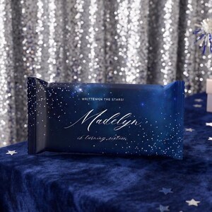 May include: A rectangular candy bar wrapper featuring a dark blue galaxy print with white stars. The text reads WRITTEN IN THE STARS! Madelyn is turning sixteen. The design includes a gradient from deep navy to lighter blue with white speckles.