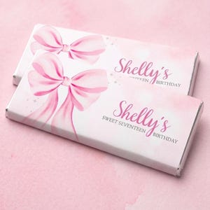 Könnte beinhalten: Zwei rechteckige Schokoriegelverpackungen mit einem rosa Schleifendesign und dem Text "Shelly's Sweet Seventeen Birthday". Die Verpackungen sind weiß mit einem rosa Aquarell-Hintergrund.
