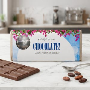 Puede incluir: Una barra de chocolate con un envoltorio decorativo que presenta una bola de discoteca, flores rosas y la palabra "CHOCOLATE!". También se muestra el texto "LUVENA TWENTY SIX BIRTHDAY". Una barra de chocolate parcialmente visible y granos de cacao en primer plano.