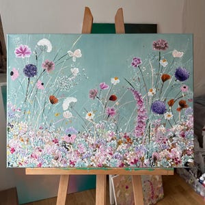 May include: An original floral painting on canvas. The artwork features a field of colorful flowers in shades of pink, purple, white, and orange against a light blue background. The painting is displayed on a wooden easel.