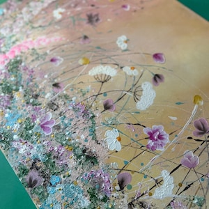 May include: An abstract painting featuring a field of flowers in shades of pink, white, purple, and blue. The artwork has a textured surface with splatters of paint and delicate lines, set against a warm, golden background.