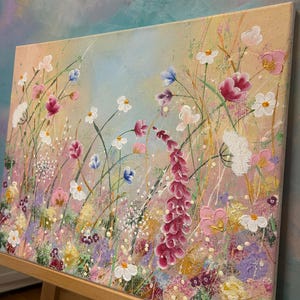 May include: An original floral painting on canvas. The artwork features a vibrant meadow of wildflowers in shades of pink, white, purple, and yellow, with a light blue and peach background. The painting is displayed on a wooden easel.