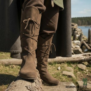 Op de afbeelding: Bruine kniehoge laarzen van suède met kruislings veters en strikdetails. De laarzen hebben een spitse neus en maken deel uit van een middeleeuws of renaissance kostuum. De laarzen worden buiten op een boomstam getoond.