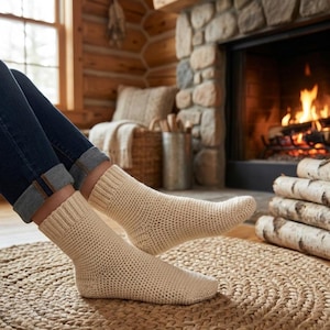 May include: Cream-colored, crocheted socks are worn by a person sitting on a woven rug. The socks are thick and textured, with a ribbed cuff. The background includes a fireplace with a burning fire, logs, and a rustic interior.