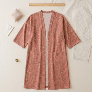 May include: A dusty rose open-front cardigan featuring a small-scale floral pattern, elbow-length sleeves, and two front patch pockets. The garment is displayed alongside a wooden ruler, a small pincushion, a tailor's chalk, and paper patterns.