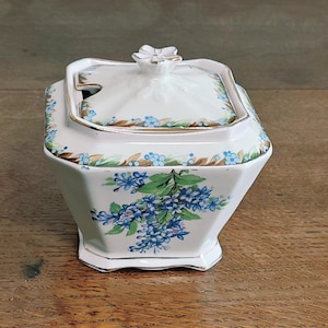 May include: A white ceramic sugar bowl with a lid, decorated with blue floral patterns and green leaves. The bowl has a gold trim and a flower-shaped handle. The sugar bowl is on a wooden surface.