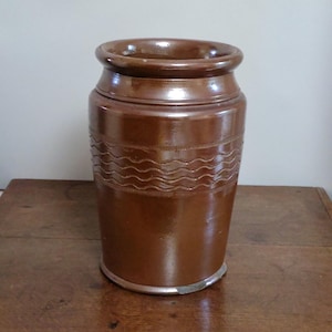 May include: A brown stoneware jar with a wide opening and a slightly flared rim. The jar features a series of wavy, horizontal lines around the middle. The jar is cylindrical and sits on a silver-coloured base. The jar is on a wooden surface.
