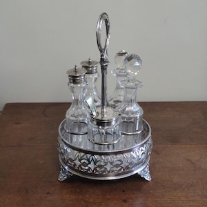 May include: A silver-plated condiment set with a decorative handle. The set includes four clear glass bottles with stoppers and a small glass container, all resting on a silver tray with ornate detailing. The tray has four feet.