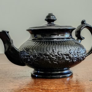 May include: A black teapot with a lid and handle. The teapot features a textured, ribbed design with floral relief patterns. The spout is curved, and the handle is arched. The teapot sits on a wooden surface.