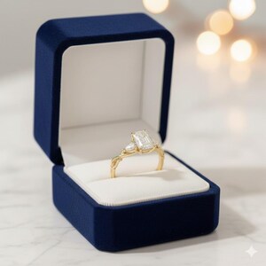 Pear diamond trio ring, radiant cut, by ExcellentJewels. Handcrafted custom jewelry ideal for engagement, proposal, or anniversary. Elegant design with timeless style and sparkling brilliance. Comfortable, durable, and personalized for daily wear or special occasions. A stunning addition to any fine jewelry collection or gift. A gold engagement ring with a rectangular center stone and two smaller side stones, set in a twisted band.