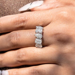 Pode incluir: Um anel de ouro branco com uma série de diamantes em várias formas, incluindo cortes de pêra, esmeralda e oval. Os diamantes são colocados próximos uns dos outros, criando uma faixa contínua de brilho. O anel é usado em uma mão com um tom de pele neutro.
