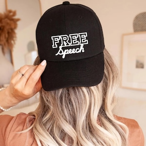 May include: Black baseball cap with the words "FREE Speech" in white block letters and script. The cap is worn by a person with long, wavy blonde hair. The person is wearing a brown top.