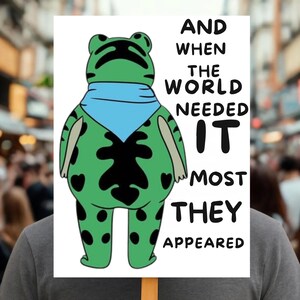May include: A sign featuring a cartoon frog with a blue scarf and black spots. The frog is green with black spots and a black design on its belly. Text on the sign reads, "AND WHEN THE WORLD NEEDED IT MOST THEY APPEARED."