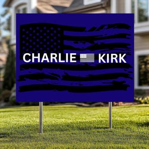 May include: A blue yard sign with the text "CHARLIE KIRK" in white, with a stylized American flag design in black and blue. The sign is mounted on two metal stakes and is displayed on a green lawn.