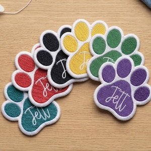 May include: A collection of colorful paw-shaped embroidered patches. Each patch features a different color, including red, black, yellow, green, teal, and purple, with a white border and a name embroidered in white script. The patches are arranged on a wooden surface.