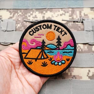 May include: A round embroidered patch with a camping scene. The patch features a tent, campfire, trees, and a sun, with the words "CUSTOM TEXT" in a curved font. The patch has a black border and is set against a camouflage background.