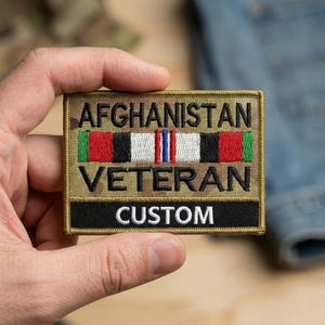 May include: A rectangular patch with the words "AFGHANISTAN VETERAN CUSTOM" in black lettering. The patch features a camouflage background and a ribbon design with red, white, black, green, and blue stripes.