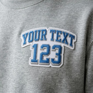 May include: A gray sweatshirt with a blue and white embroidered patch. The patch features the text "YOUR TEXT" above the numbers "123". The text and numbers are in a bold, collegiate style font.