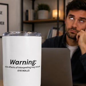 May include: A white stainless steel tumbler with a clear lid featuring the black printed text: Warning: side effects of interpreting may include EYE ROLLS. The cylindrical drinkware has a tapered base and a smooth, matte finish on the exterior surface.