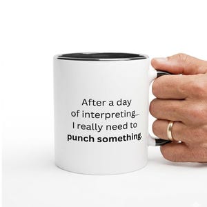 May include: White ceramic mug with a black rim and handle. The mug features the text "After a day of interpreting... I really need to punch something." A hand with a gold ring is holding the mug.
