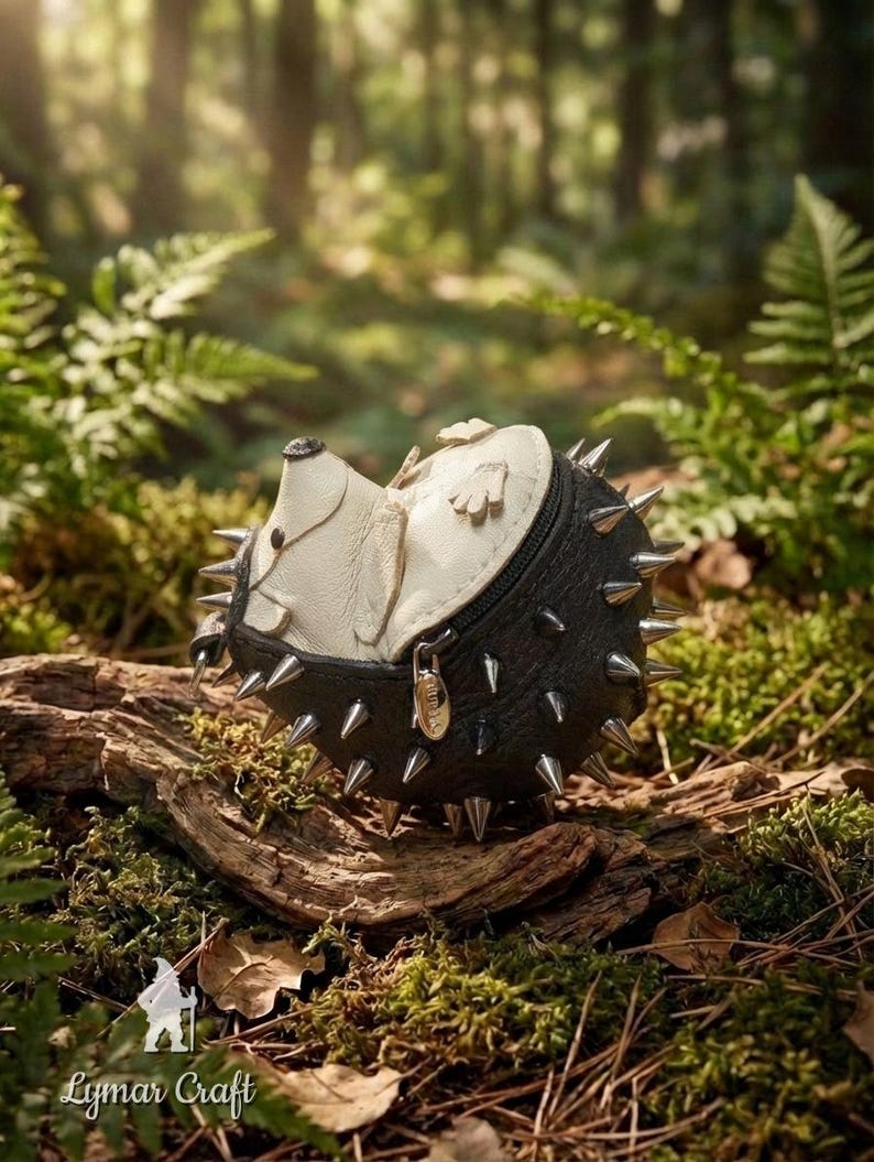 May include: A handcrafted, black and cream-colored leather hedgehog-shaped coin purse with silver-toned spikes. The purse is lying on a piece of wood in a forest setting, surrounded by green moss and ferns. The zipper has a gold-toned pull.