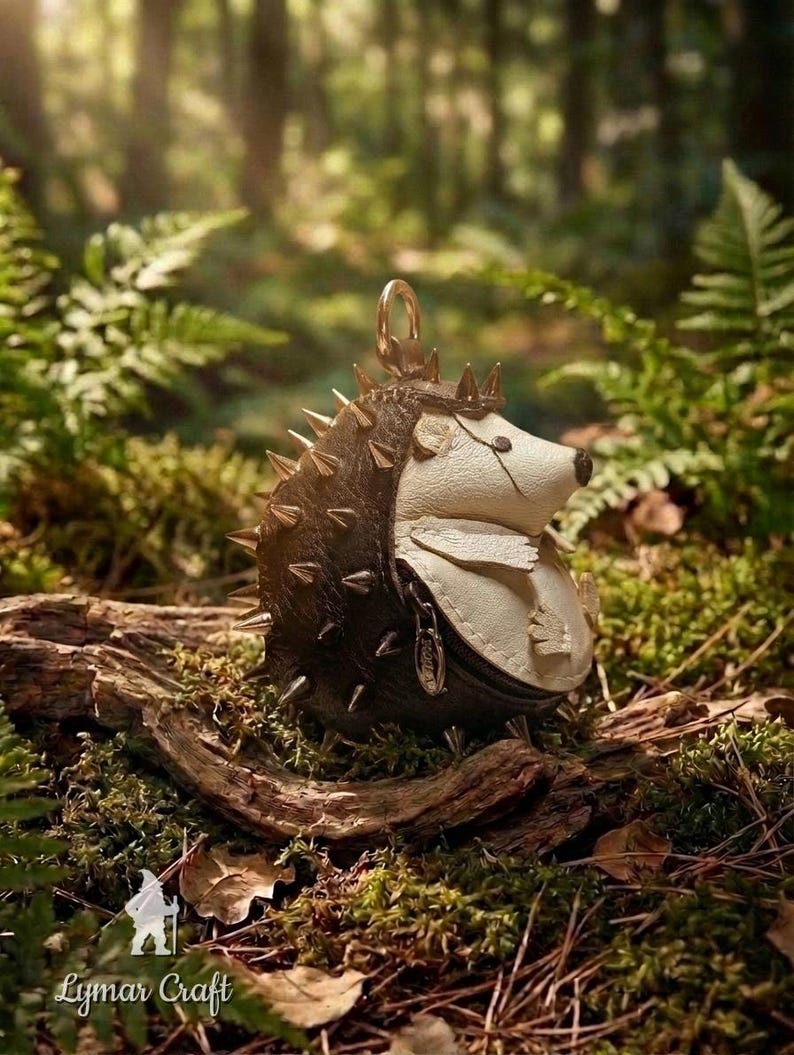 May include: A handcrafted leather hedgehog-shaped coin purse with a crown and metal spikes. The purse is black and cream-colored, with a zipper closure and a gold-tone ring for attaching to a keychain. The purse is set on a bed of moss and wood.