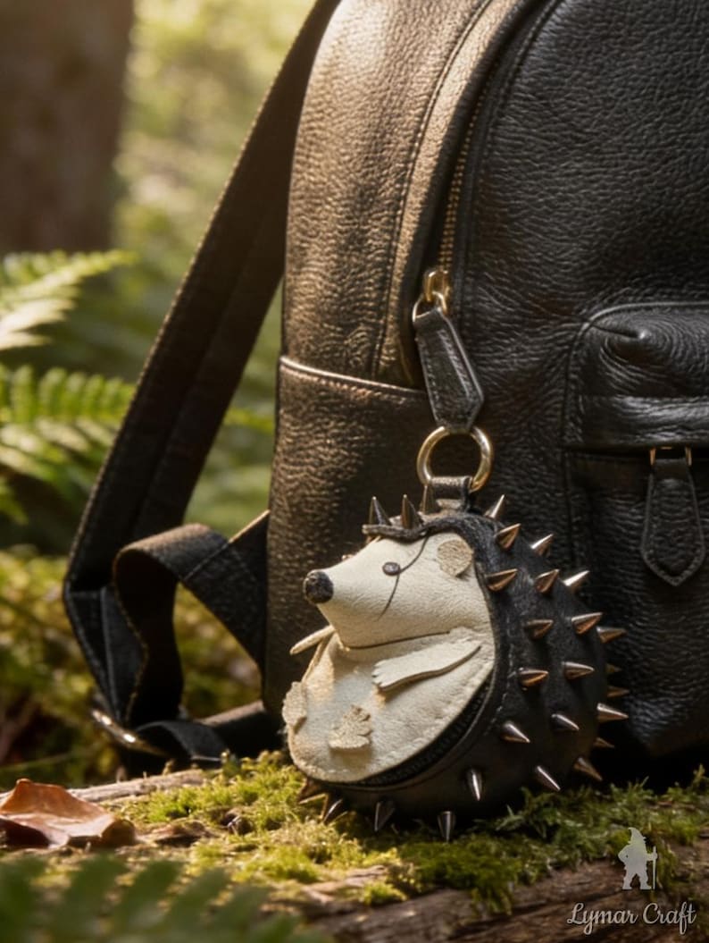 May include: A black leather backpack with a hedgehog-shaped keychain. The keychain is black and cream-colored with metal spikes. The hedgehog has a small leaf detail on its belly. The background is a natural setting with green foliage.