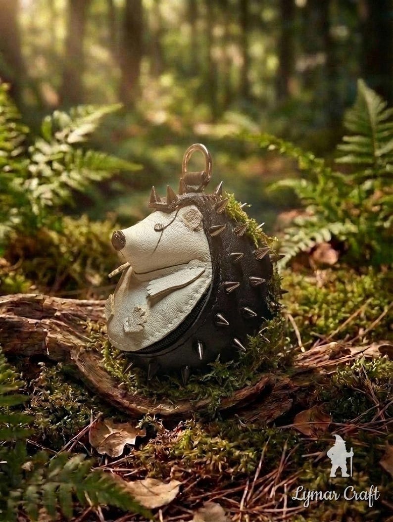 May include: A whimsical, handcrafted hedgehog-shaped coin purse. The purse features a white face, a black body with silver spikes, and a small crown. It is set against a natural forest backdrop with moss and ferns.