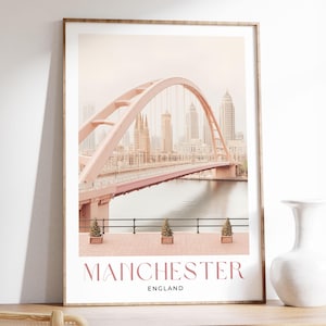 Può includere: Stampa incorniciata con un ponte rosa ad arco sull'acqua, con uno skyline sullo sfondo. La stampa presenta il testo "MANCHESTER ENGLAND" in rosa. La cornice è di colore legno chiaro.