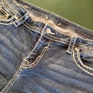 May include: Close-up of blue denim jeans with white stitching. The jeans feature a button closure, front pockets, and decorative stitching around the pockets and seams. The denim has a faded appearance.