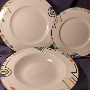 May include: A set of three white ceramic plates with a colorful geometric design. The plates include a dinner plate, a salad plate, and a soup bowl. The design features black lines, spirals, and colorful brushstroke-like shapes.