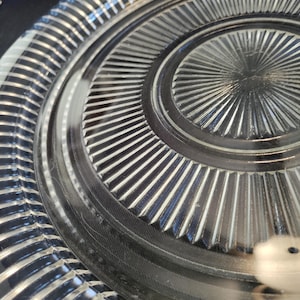 May include: Close-up of a clear glass plate with a radial design. The plate features a series of concentric circles and radiating lines, creating a textured surface. The edges have a ribbed pattern.