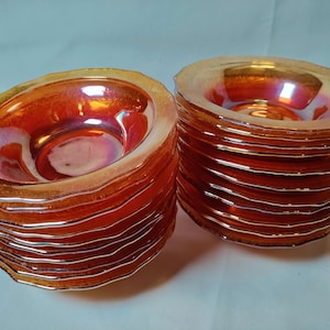 May include: Two stacks of round glass bowls featuring a vibrant orange iridescent finish. Each bowl has a scalloped edge and a smooth, reflective surface that catches the light, creating a warm, metallic glow across the stacked set.