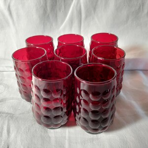 May include: Set of eight ruby red drinking glasses. Each glass has a textured, bubble-like pattern. The glasses are cylindrical with a wide opening and a thick base. The glasses are arranged in a cluster, showcasing their vibrant color and design.