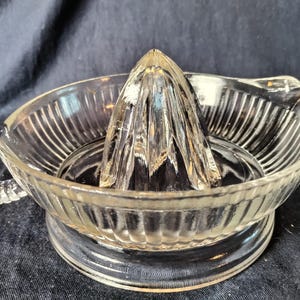 May include: Clear glass citrus juicer with a ribbed bowl, a handle, and a central reamer. The juicer is designed for easy citrus fruit juicing. The glass has a vintage look.