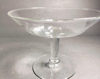 May include: A clear glass pedestal bowl with a wide, shallow bowl and a tall, slender stem. The bowl is perfect for serving desserts, appetizers, or other small dishes.