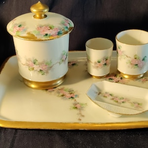 May include: A vintage porcelain vanity set featuring a covered jar, two cups, and a small dish on a rectangular tray. The set is white with pink floral designs, gold accents, and a gold rim.