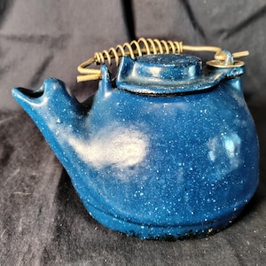 May include: A small, blue enamelware teapot with white speckles. It has a spout, a lid, and a wire handle with a coiled grip. The teapot is designed for camping or outdoor use.