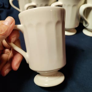 May include: A set of off-white ceramic mugs, each featuring a vertical ribbed pattern along the body and a pedestal base. The mugs have a smooth, glossy finish and a curved handle attached to the side, with one mug held in the foreground.