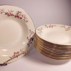 May include: A stack of 12 white ceramic soup bowls with a gold rim and a pink floral design. The bowls are stacked on a white surface.