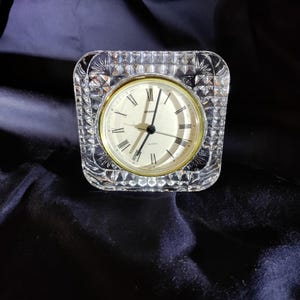 May include: A clear, square quartz clock with a gold-colored bezel and Roman numerals. The clock face is cream-colored, and the crystal casing has a diamond-cut pattern. The clock is a decorative item.