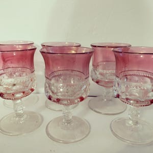May include: Six vintage wine glasses with a clear, ornate base and a vibrant pink rim. The glasses have a classic, elegant design, perfect for serving wine or other beverages. The stemware is arranged on a white surface.