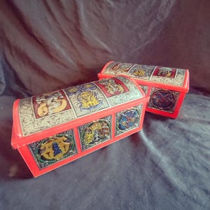 May include: Two red toy chests with arched lids. Each chest features decorative panels with heraldic designs in gold, blue, and other colors. The chests are set against a dark, textured background.