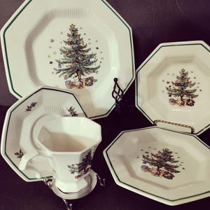 May include: A collection of white octagonal Christmas-themed dinnerware. The plates and teacup feature a green border and a Christmas tree design with gifts. The teacup sits on a silver stand.