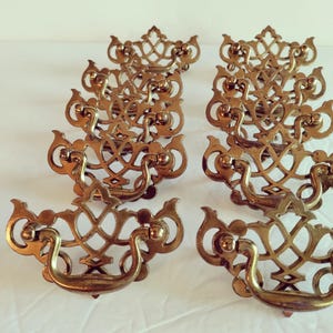 May include: A collection of antique brass drawer pulls. Each pull features an ornate, openwork design with a curved handle and decorative details. The brass has a warm, aged patina, suggesting vintage craftsmanship. These pulls are likely for furniture restoration.