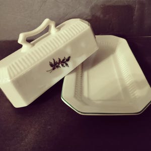 May include: White ceramic butter dish with a lid and handle. The lid features a sprig of berries and leaves. The dish has a rectangular shape with a green trim around the edge. The dish is open, revealing the interior.