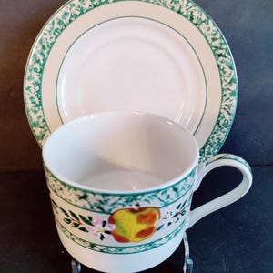 May include: A white teacup and saucer with a green and white floral pattern. The cup features a painted apple with pink and yellow flowers.